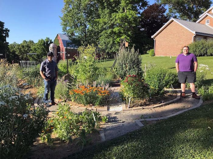 Two men stand beside a collection of plants in the sun